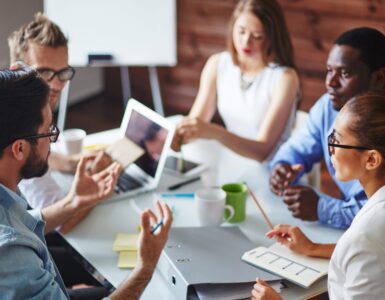 Multiple coworkers sit at a desk in an office while talking, with one man gesturing during the discussion.