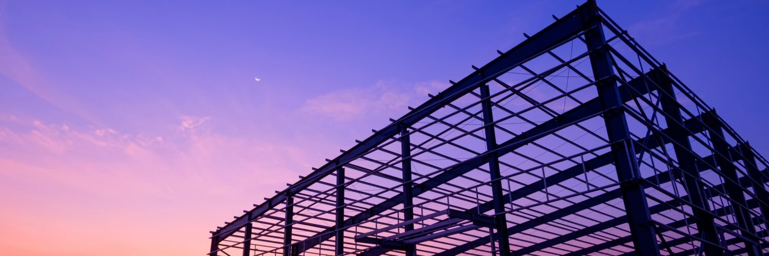 The steel structure of a partially built industrial warehouse, with the sunset behind the framework.