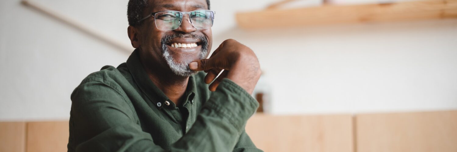 A smiling man wearing glasses and a green shirt sits at a table, resting his chin on his hand in a room with wooden shelves.