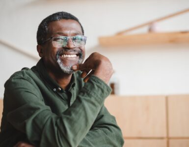 A smiling man wearing glasses and a green shirt sits at a table, resting his chin on his hand in a room with wooden shelves.