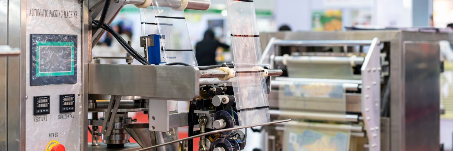 A pouch packaging machine feeding film from rollers, showing sealing components and panel on a factory floor.