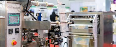 A pouch packaging machine feeding film from rollers, showing sealing components and panel on a factory floor.