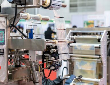 A pouch packaging machine feeding film from rollers, showing sealing components and panel on a factory floor.