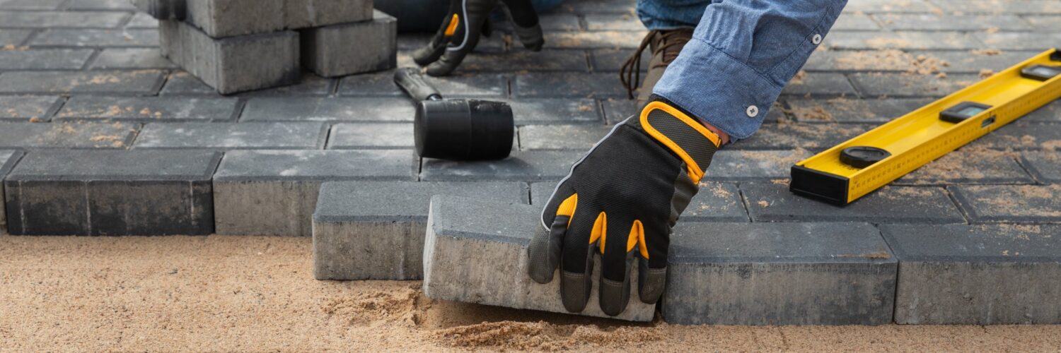 A close-up of a person wearing black and yellow work gloves while laying brick pavers on top of compacted sand.
