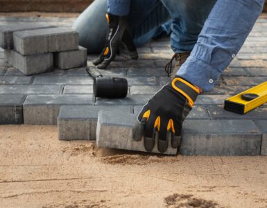 A close-up of a person wearing black and yellow work gloves while laying brick pavers on top of compacted sand.
