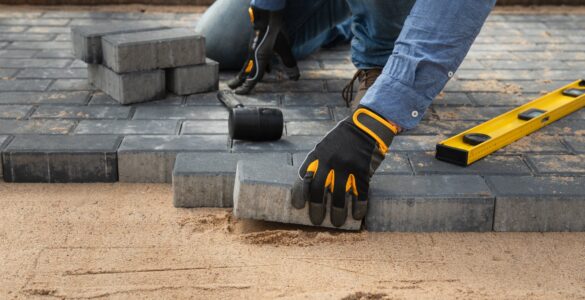 A close-up of a person wearing black and yellow work gloves while laying brick pavers on top of compacted sand.
