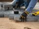 A close-up of a person wearing black and yellow work gloves while laying brick pavers on top of compacted sand.