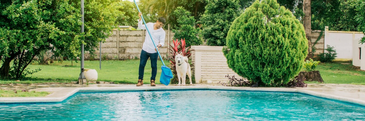 A man holding a long-handled net over a swimming pool in a backyard. There is a dog standing next to the man.