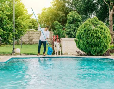 A man holding a long-handled net over a swimming pool in a backyard. There is a dog standing next to the man.
